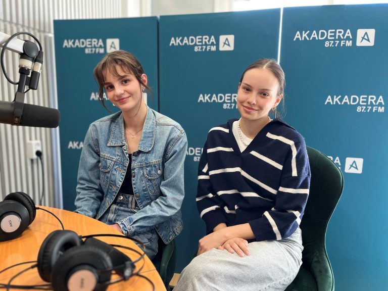 Dwie młode kobiety siedzące w studiu radiowym Radia Akadera. W tle widoczne niebieskie ścianki z logo rozgłośni „AKADERA 87.7 FM”. Po lewej stronie znajduje się mikrofon radiowy i leżące na stole słuchawki. Kobieta po lewej ma na sobie dżinsową kurtkę i czarny top, a jej włosy są związane w luźny kucyk. Kobieta po prawej ubrana jest w granatowy sweter w białe paski i jasnoszarą spódnicę, jej włosy są zaczesane do tyłu. Obie patrzą w stronę obiektywu i uśmiechają się delikatnie.