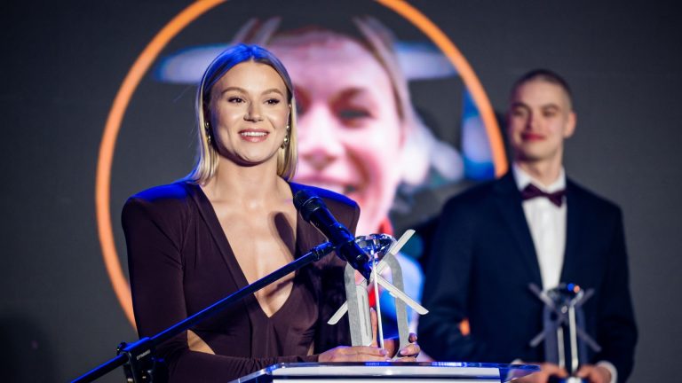 Natalia Maliszewska uśmiecha się, stojąc przy mikrofonie i trzymając statuetkę Kryształowej Płozy. W tle rozmyty portret na ekranie. Obok niej młody mężczyzna w garniturze z muchą i drugą statuetką w dłoniach.