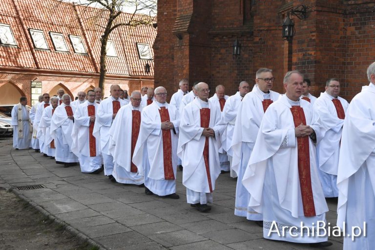 Grupa kilkudziesięciu księży ubranych w białe alby z czerwonymi stułami uczestniczy w uroczystej procesji przed katedrą w Białymstoku z okazji Wielkiego Czwartku. W tle widoczne ceglane mury świątyni i budynki Arkad Katedralnych.