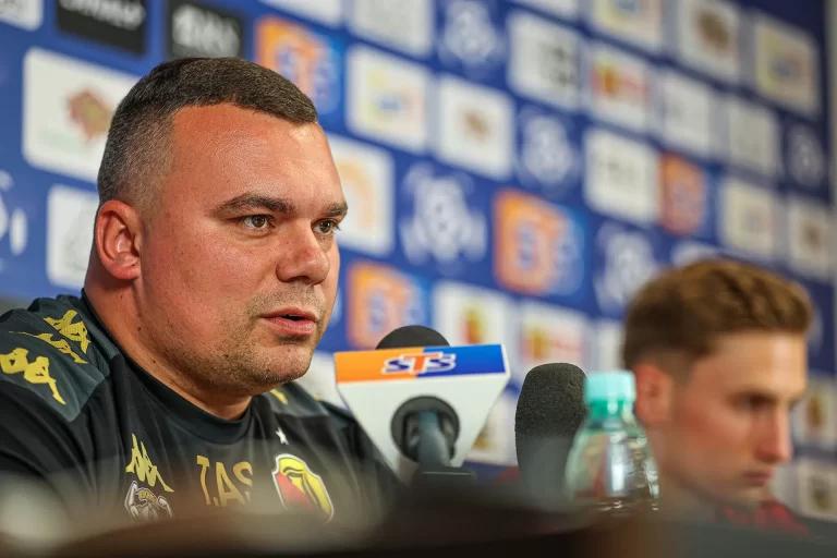 Adrian Siemieniec, trener Jagiellonii Białystok, w czarnej koszulce z herbem klubu i logotypami sponsorów, mówi do mikrofonów podczas konferencji prasowej. W tle ścianka z logotypami medialnymi i sponsorskimi. Obok niego rozmyta sylwetka piłkarza.