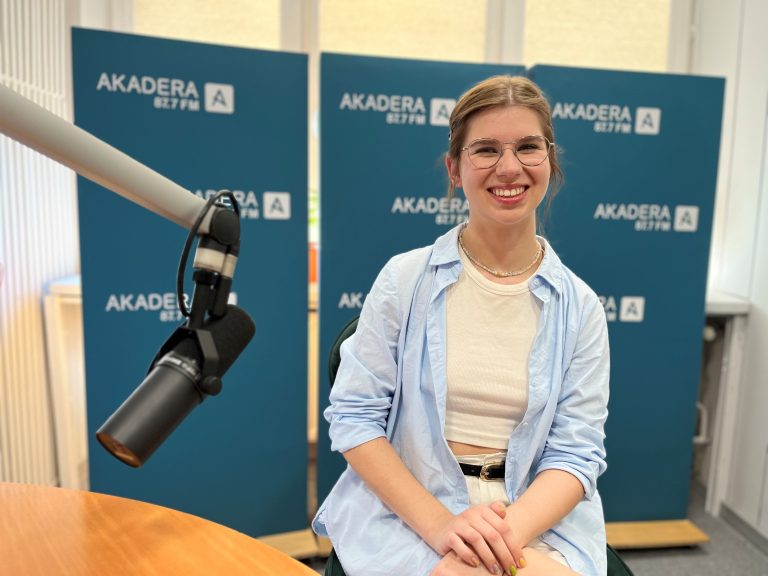 Młoda kobieta z długimi, jasnymi włosami siedzi przy mikrofonie w studiu Radia AKADERA. Ubrana w jasny sweter, z uśmiechem patrzy w stronę rozmówcy. W tle ścianka z logo AKADERA 87.7 FM oraz profesjonalne wyposażenie radiowe.