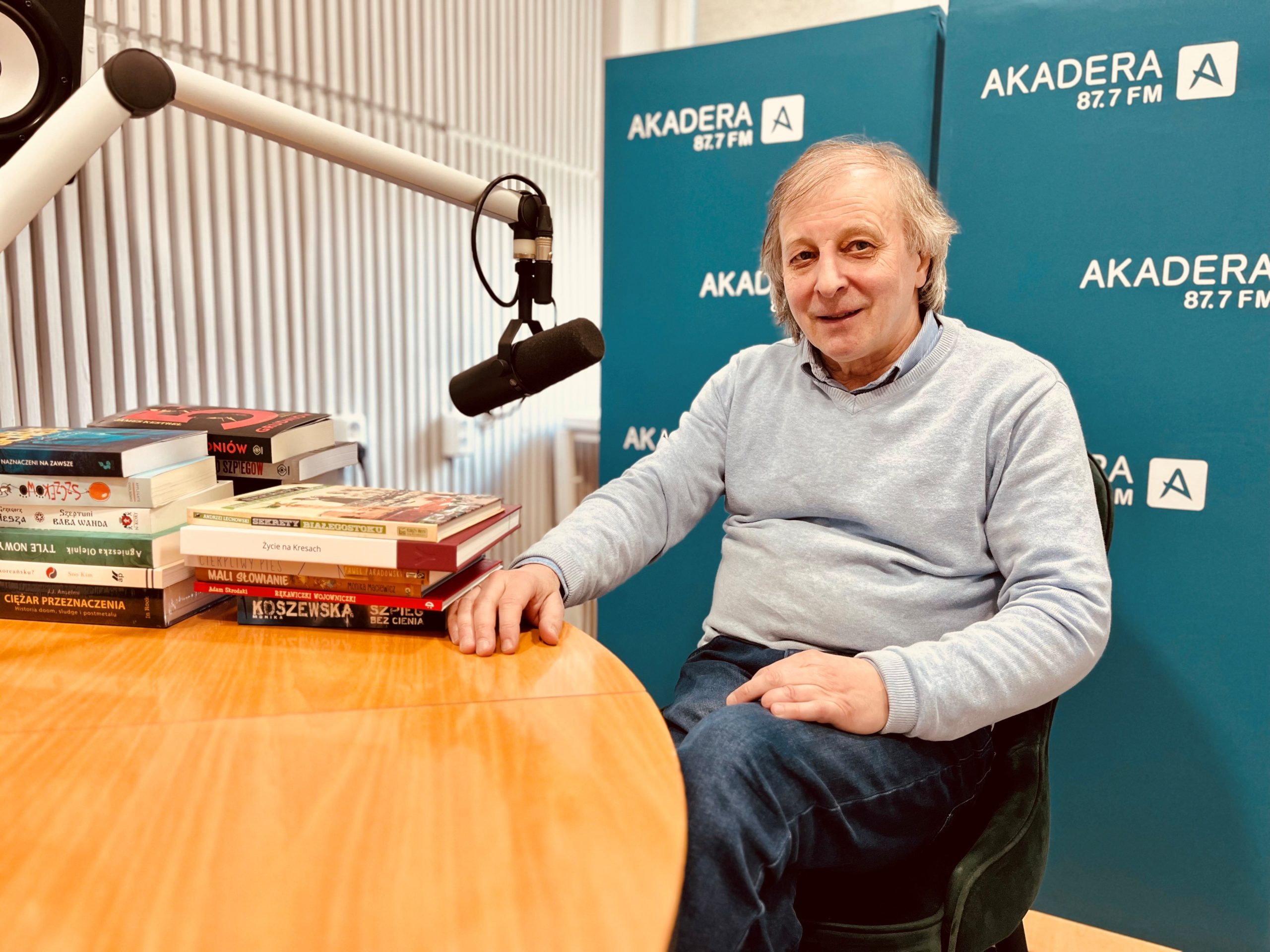 Andrzej Kalinowski, organizator Targów Książki w Białymstoku, siedzi przy stole w radiowym studiu Radia Akadera. Przed nim mikrofon oraz stos kolorowych książek. W tle niebieskie ścianki z logo Akadera 87.7 FM.