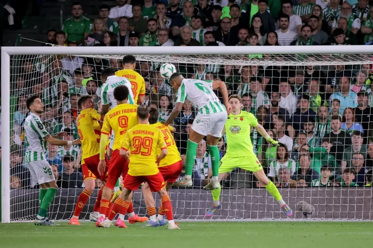 Piłkarze Realu Betis i Jagiellonii Białystok w akcji podbramkowej – zawodnik Betisu wyskakuje do główki, bramkarz Jagiellonii przygotowany do interwencji. W tle tłum kibiców w zielono-białych barwach zasiadających na trybunach stadionu Benito Villamarín w Sewilli.