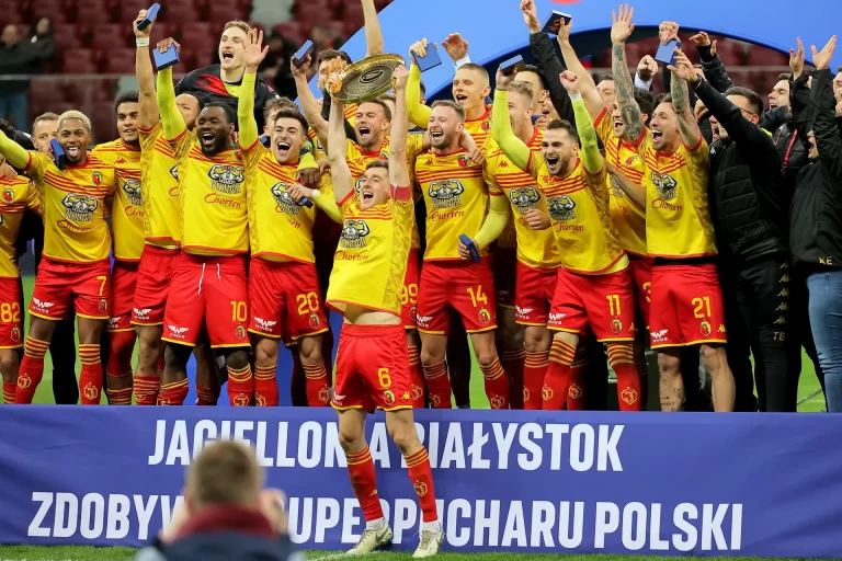Piłkarze Jagiellonii Białystok w żółto-czerwonych strojach świętują zdobycie Superpucharu Polski. Na środku zawodnik z numerem 6 unosi trofeum w górę, wokół niego zespół wiwatuje z uniesionymi rękami. W tle widoczne są trybuny Stadionu Narodowego.