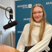 Młoda kobieta z długimi blond włosami i okularami, ubrana w jasny sweter, siedzi przy stole radiowym w studiu Radia AKADERA. Obok niej mikrofon studyjny. W tle ścianka z logo AKADERA 87.7 FM.