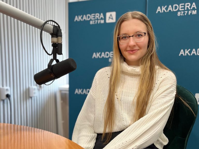 Młoda kobieta z długimi blond włosami i okularami, ubrana w jasny sweter, siedzi przy stole radiowym w studiu Radia AKADERA. Obok niej mikrofon studyjny. W tle ścianka z logo AKADERA 87.7 FM.