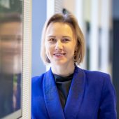 Dr Martyna Faustyna Zaniewska, dyrektorka Białostockiego Ośrodka Kultury, uśmiechnięta, ubrana w niebieską marynarkę, stoi przy ścianie z ekranami w jasnym wnętrzu.