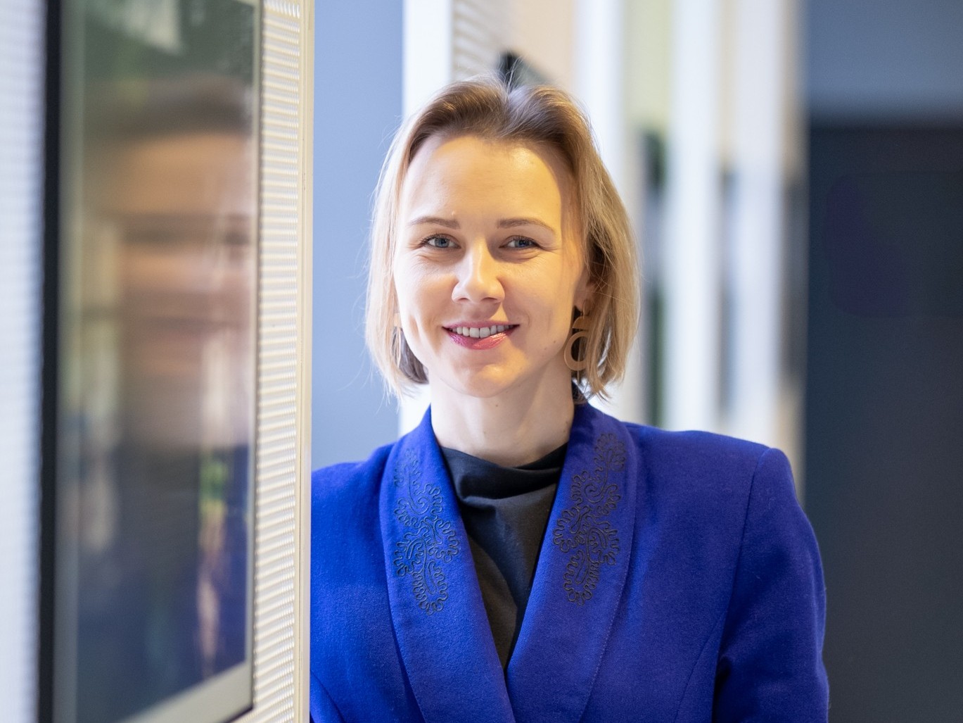 Dr Martyna Faustyna Zaniewska, dyrektorka Białostockiego Ośrodka Kultury, uśmiechnięta, ubrana w niebieską marynarkę, stoi przy ścianie z ekranami w jasnym wnętrzu.