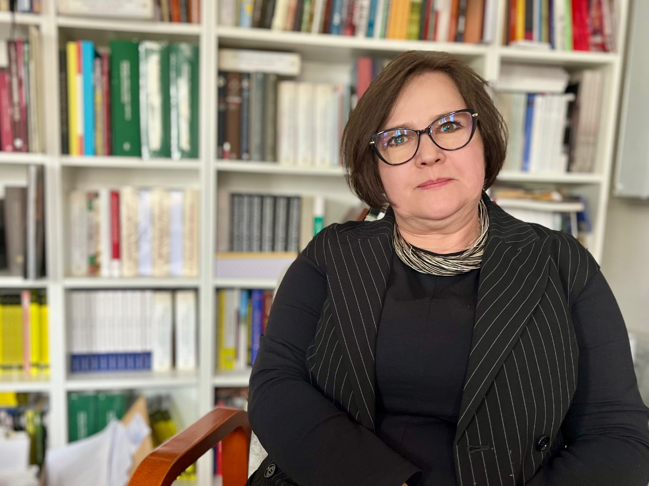 Kobieta w ciemnych okularach i eleganckim stroju siedzi na tle regału pełnego książek; to prof. Marzena Liedke z Uniwersytetu w Białymstoku.