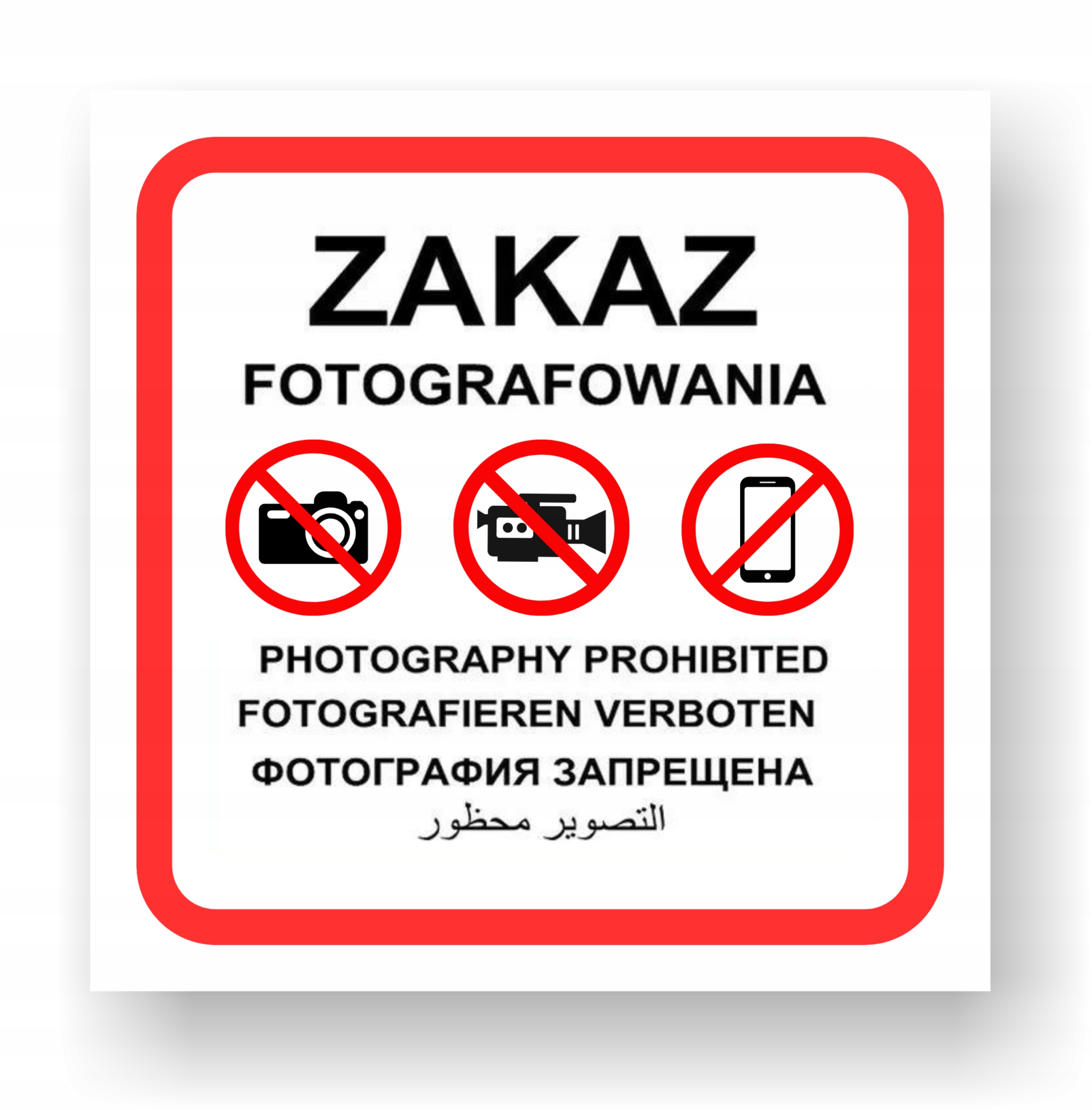 Kwadratowa tablica z czerwonym obramowaniem i czarnym napisem „Zakaz fotografowania”. Pod nim znajdują się trzy przekreślone piktogramy: aparatu fotograficznego, kamery i telefonu komórkowego. Niżej znajduje się ten sam komunikat przetłumaczony na języki: angielski, niemiecki, rosyjski i arabski.
