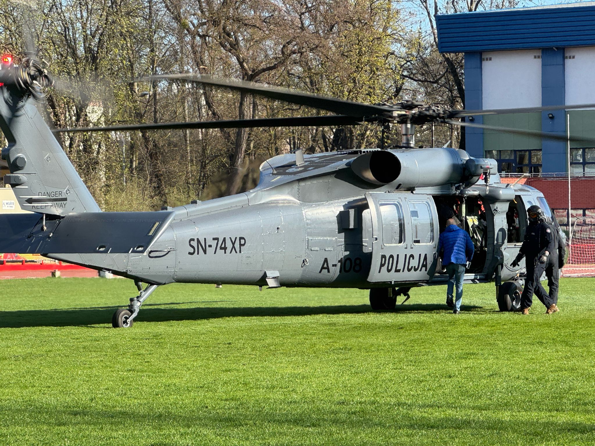 Policyjny śmigłowiec Black Hawk z otwartymi drzwiami, gotowy do startu z trawiastych obiektów stadionu w Białymstoku. Obok maszyny stoją ratownicy i personel. Helikopter jest oznaczony napisem „Policja” i numerem SN-74XP.