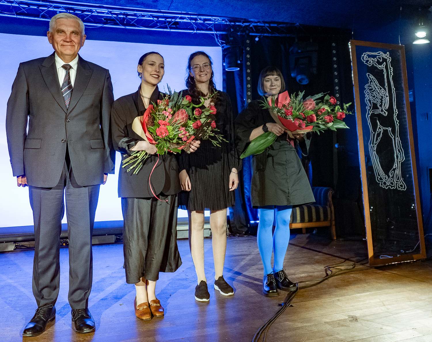 Prezydent Białegostoku Tadeusz Truskolaski oraz laureatki Nagrody Literackiej Kazaneckiego 2025 stoją na scenie z bukietami kwiatów, na tle niebieskiego światła i dekoracji.