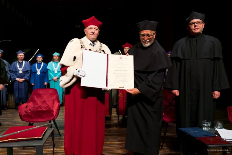 Rektor Uniwersytetu w Białymstoku prof. Mariusz Popławski i prof. Piotr Hofmański prezentują dyplom nadania tytułu doktora honoris causa podczas uroczystości akademickiej. Obaj ubrani są w togi – rektor w czerwonej z gronostajową peleryną, prof. Hofmański w czarnej todze z biretem. W tle stoją inni uczestnicy uroczystości w togach w różnych kolorach.