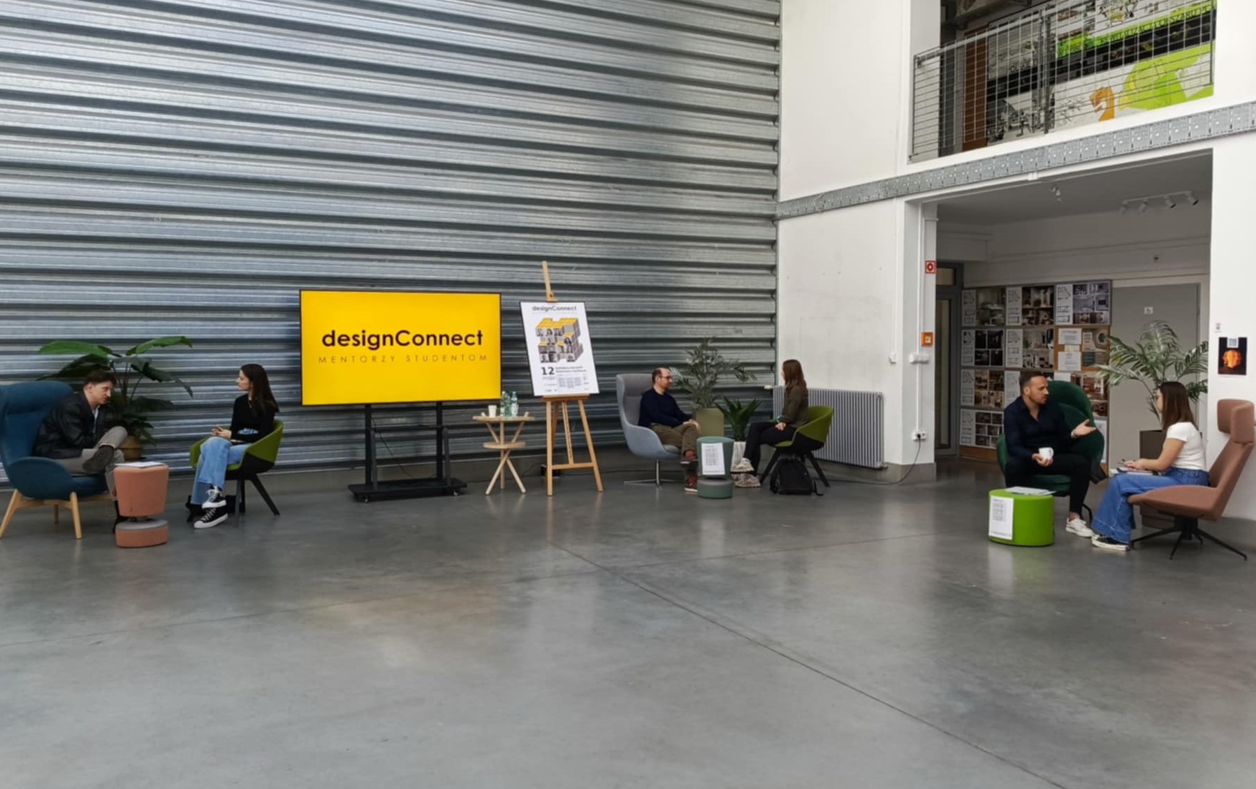 Na zdjęciu hala na Wydziale Architektury PB, na niej duzy ekran z napisem na żółtym tle DESIGN CONNECT. Przy stolikach siedzą mentorzy oraz studenci, trwają rozmowy i porady.