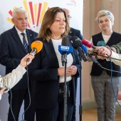 Kobieta w ciemnym garniturze udziela wypowiedzi podczas konferencji prasowej, stojąc przy mikrofonach różnych mediów. W tle widoczni są inni uczestnicy wydarzenia, w tym mężczyźni i kobiety w strojach formalnych. Na ścianie znajduje się baner z logo miasta Białystok.