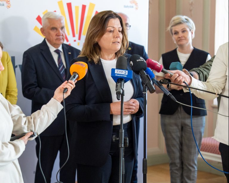 Kobieta w ciemnym garniturze udziela wypowiedzi podczas konferencji prasowej, stojąc przy mikrofonach różnych mediów. W tle widoczni są inni uczestnicy wydarzenia, w tym mężczyźni i kobiety w strojach formalnych. Na ścianie znajduje się baner z logo miasta Białystok.