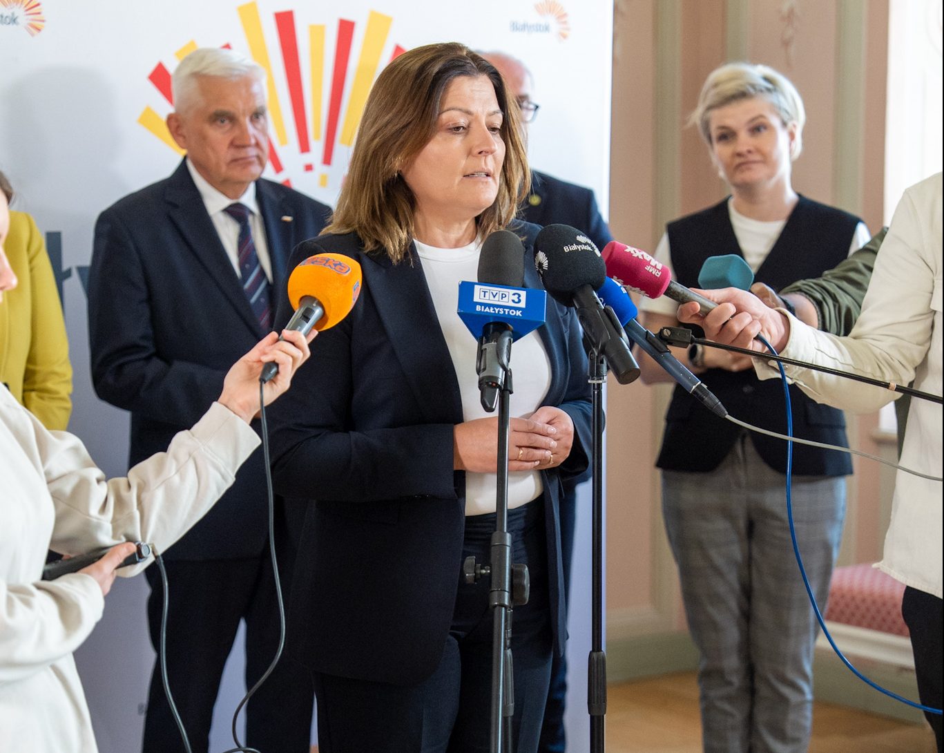 Kobieta w ciemnym garniturze udziela wypowiedzi podczas konferencji prasowej, stojąc przy mikrofonach różnych mediów. W tle widoczni są inni uczestnicy wydarzenia, w tym mężczyźni i kobiety w strojach formalnych. Na ścianie znajduje się baner z logo miasta Białystok.