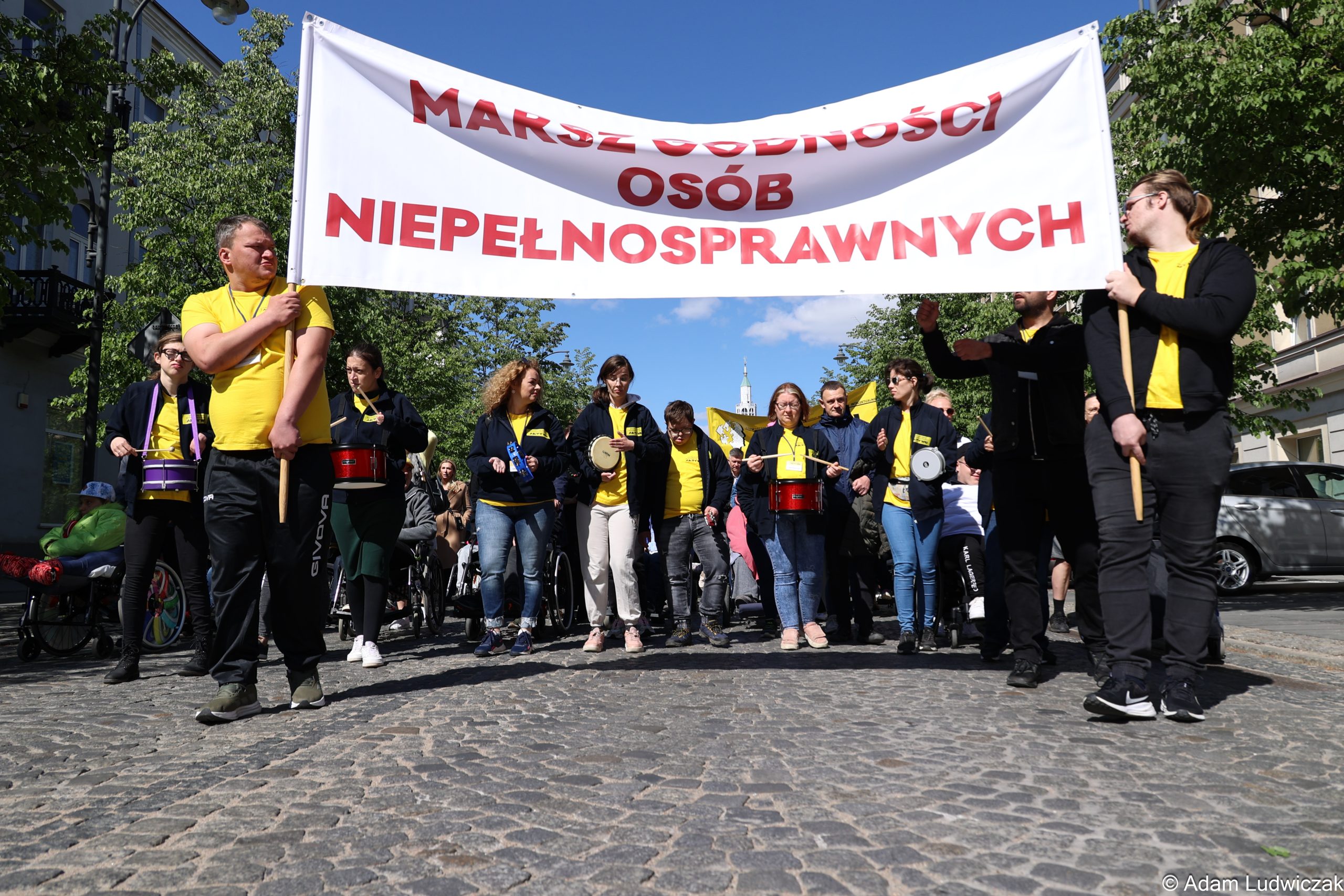 Duży czerwony transparent z białym napisem „MARSZ OSÓB NIEPEŁNOSPRAWNYCH” niesiony przez uczestników. W tle flaga Polski i kamienice.