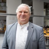 Dr inż. arch. Adam Jakimowicz, ubrany w szarą marynarkę i białą koszulę, uśmiecha się do obiektywu. W tle widać przestronną salę z ustawionymi rzędami drewnianych krzeseł i stołów oraz studentów pracujących nad projektami.