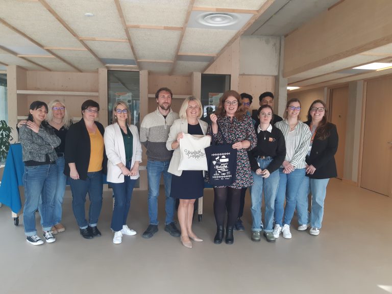 Grupa uśmiechniętych osób pozująca do zdjęcia w przestronnym pomieszczeniu. Prof. Joanna Mystkowska stoi w centrum grupy, trzymając pamiątkową torbę.