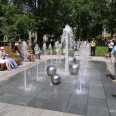 Ludzie odpoczywający na nowoczesnych ławkach wokół fontanny na Skwerze im. Pawła Adamowicza w Białymstoku. Fontanna z wodotryskami otoczona kulistymi elementami dekoracyjnymi, w tle drzewa, przechodnie i namioty eventowe.
