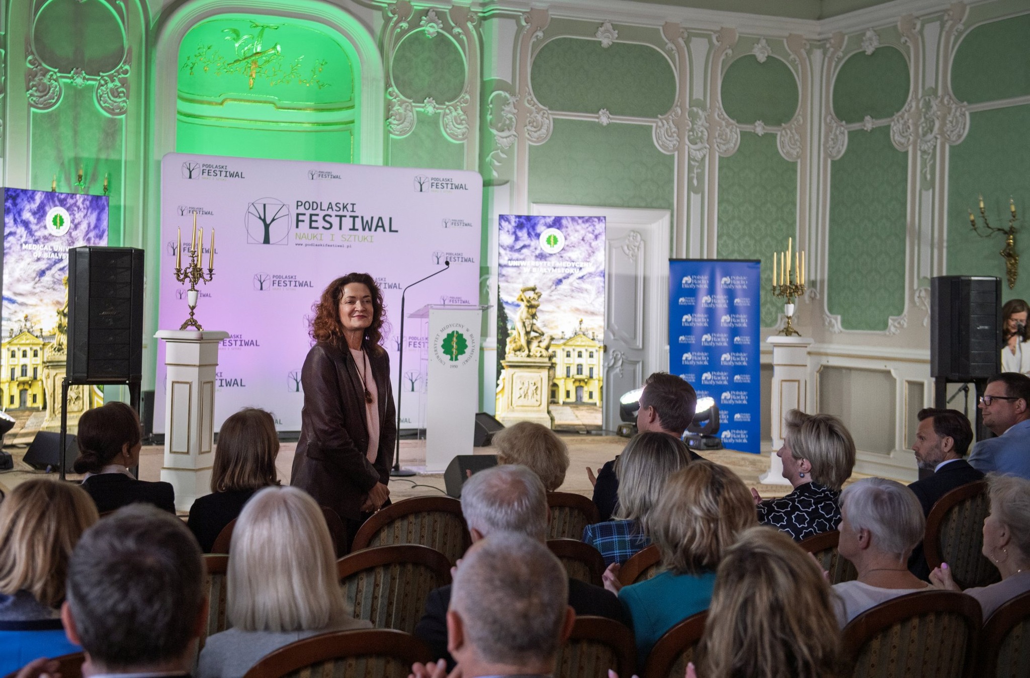 Prof. Marta Kosior-Kazberuk, Rektor Politechniki Białostockiej, stoi uśmiechnięta przed publicznością podczas inauguracji Podlaskiego Festiwalu Nauki i Sztuki w eleganckiej sali z zielonymi ścianami i sceną udekorowaną banerami festiwalowymi.