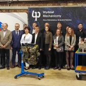 Na tle ścianki z napisem wydzial mechaniczny politechniki bialostockiej stoja elegancko ubrani przedstawciele swiata nauki i biznesu. Przed nimi dwa przekroje silnikow, majacych sluzyc jako pomoze dydaktyczne