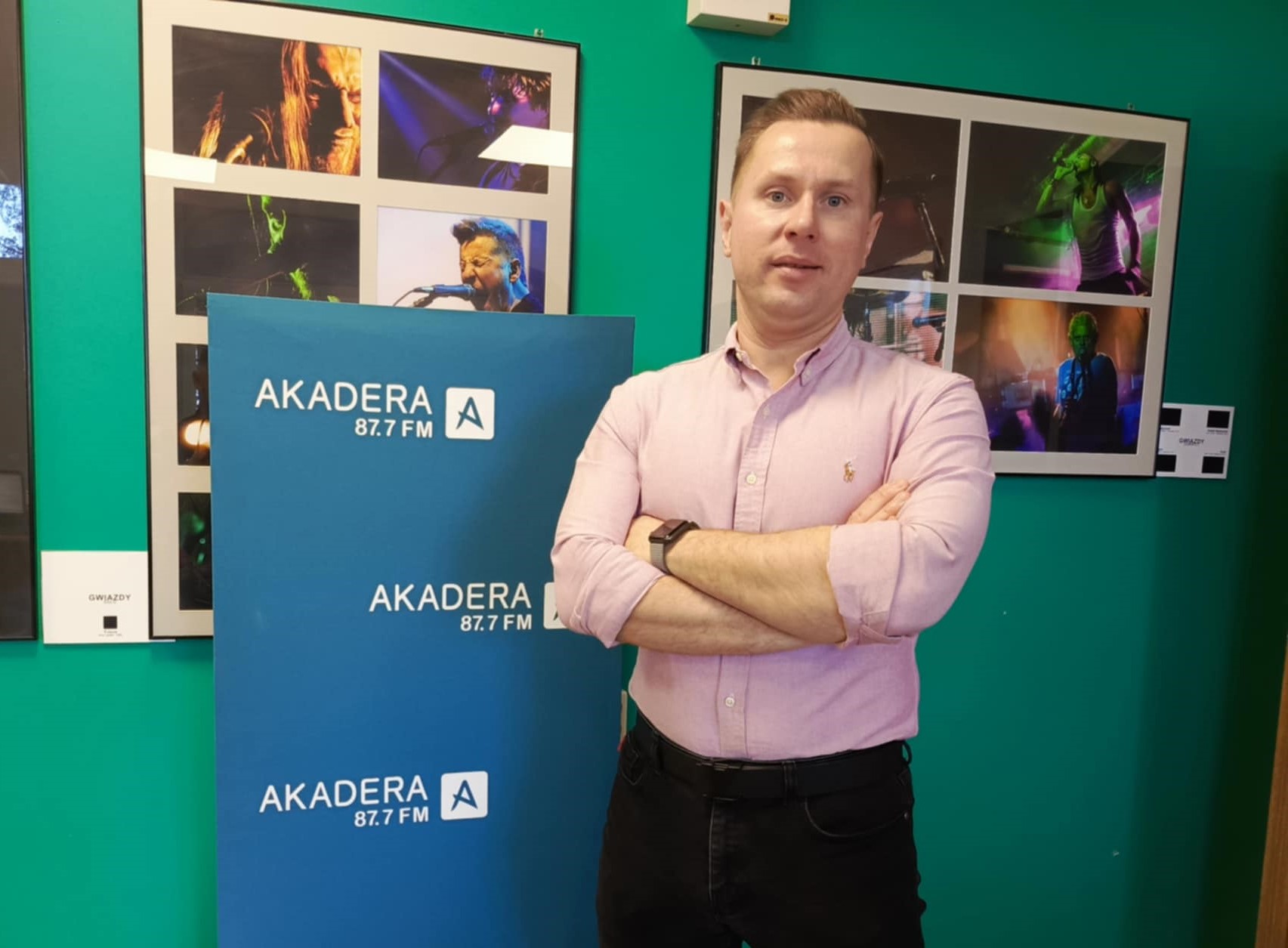 Na zdjeciu mezczyzna ubrany w rozowa koszule, elegancki, na tle niebieskiej planszy Radio Akadera i zdjec zespolow na koncertach.