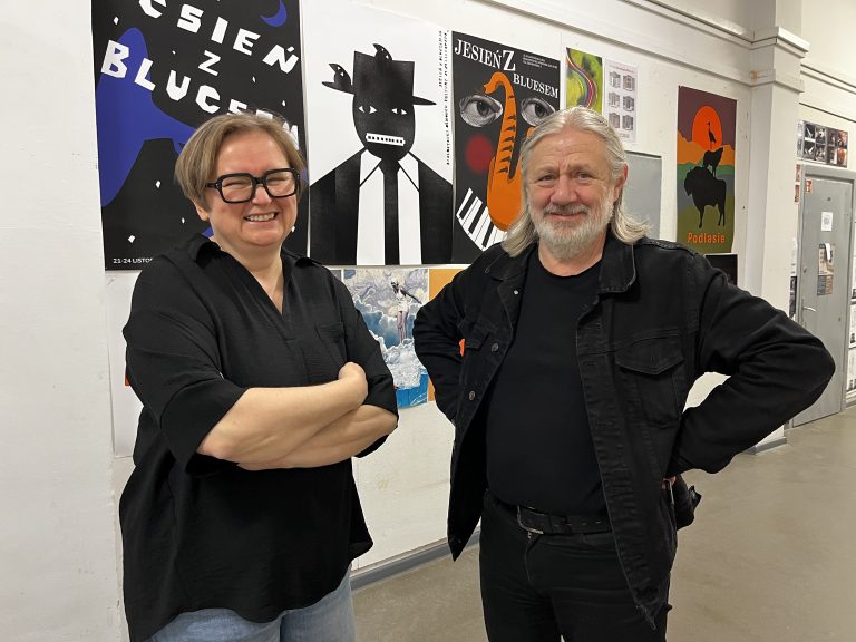 Dr hab. Andrzej Dworakowski i mgr Katarzyna Zawłocka stojący w korytarzu Wydziału Architektury Politechniki Białostockiej, na tle ściany z pracami studentów grafiki – plakatami artystycznymi.