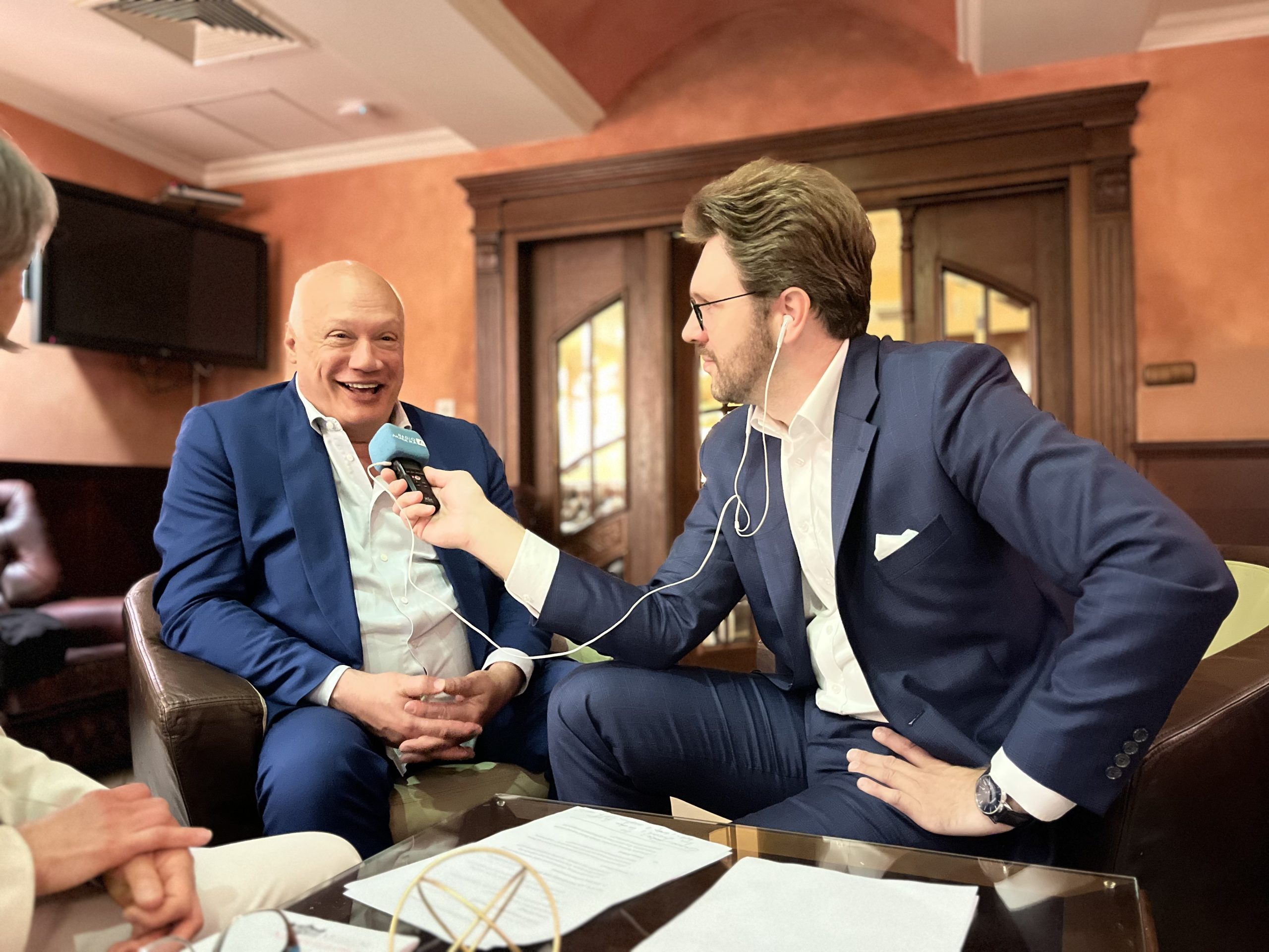 Pisarz Éric-Emmanuel Schmitt uśmiechnięty podczas wywiadu z dziennikarzem Adamem Jakuciem w eleganckim wnętrzu, obaj ubrani w granatowe garnitury, trzymają mikrofon z logo Radia Akadera.