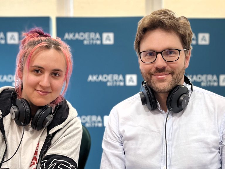 Uśmiechnięta kobieta z różowymi włosami i mężczyzna w okularach siedzą obok siebie w studiu Radia Akadera. Oboje mają słuchawki radiowe na szyi, w tle plansze z logo Akadera 87,7 FM.