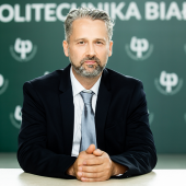 Dr hab. inż. Jarosław Szusta w garniturze, siedzący przy stole na tle logotypu Politechniki Białostockiej, patrzy w obiektyw.