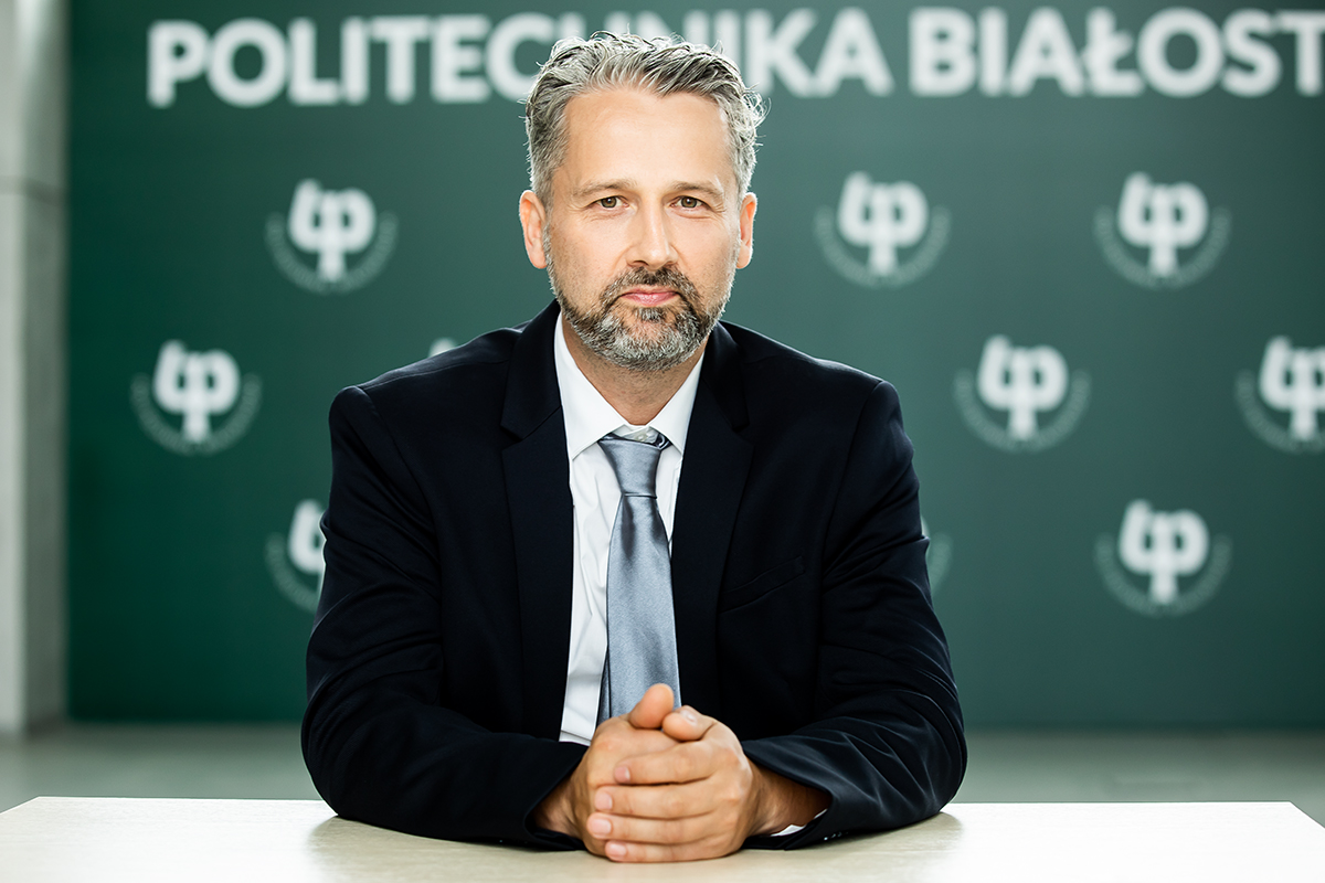 Dr hab. inż. Jarosław Szusta w garniturze, siedzący przy stole na tle logotypu Politechniki Białostockiej, patrzy w obiektyw.