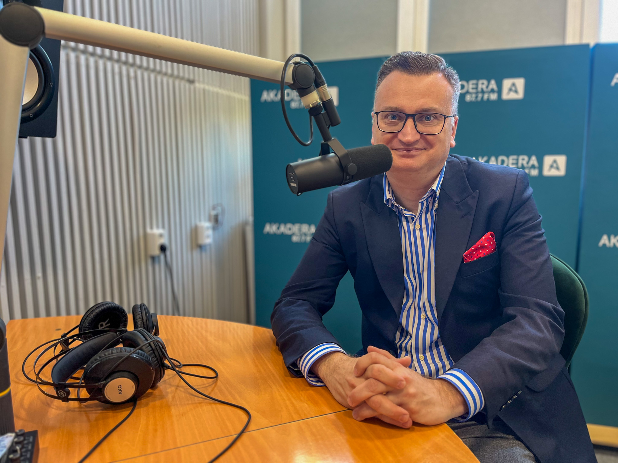 Wiceprezydent Rafał Rudnicki siedzi przy mikrofonie w studiu Radia Akadera; ma granatową marynarkę, koszulę w niebiesko-białe pasy i czerwony poszetka, przed nim leżą studyjne słuchawki.