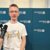 Młody mężczyzna w okularach i białej koszulce z napisem „Elektryk zawsze pozytywnie naładowany” siedzi w studiu Radia Akadera, przed mikrofonem. W tle niebieskie ścianki z logo Akadera 87.7 FM.