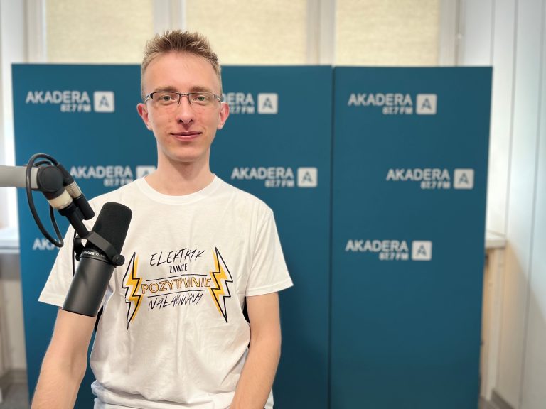 Młody mężczyzna w okularach i białej koszulce z napisem „Elektryk zawsze pozytywnie naładowany” siedzi w studiu Radia Akadera, przed mikrofonem. W tle niebieskie ścianki z logo Akadera 87.7 FM.