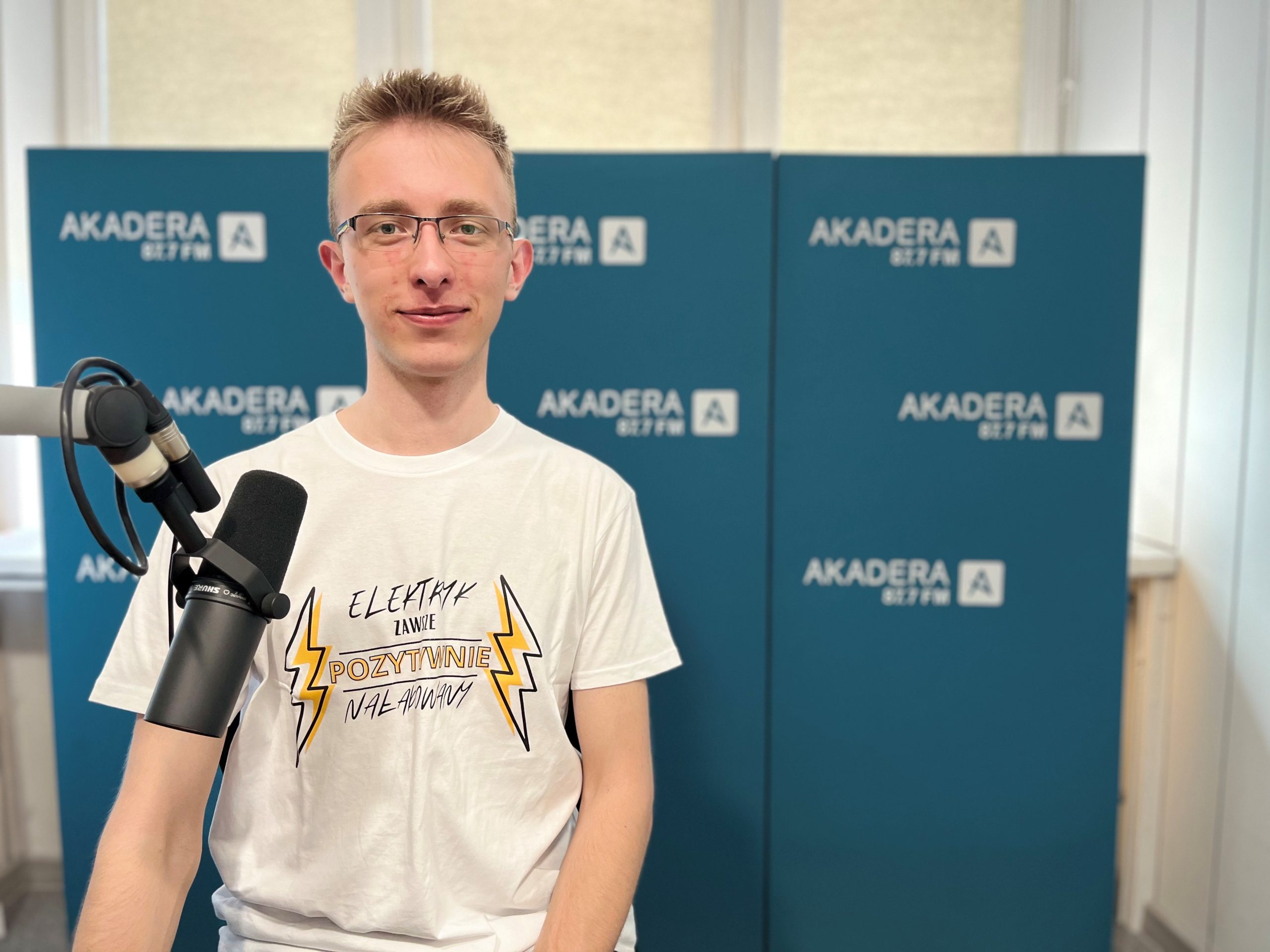 Młody mężczyzna w okularach i białej koszulce z napisem „Elektryk zawsze pozytywnie naładowany” siedzi w studiu Radia Akadera, przed mikrofonem. W tle niebieskie ścianki z logo Akadera 87.7 FM.