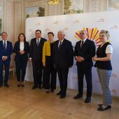 Grupa siedmiorga osób w eleganckich strojach w tym Prezydent Białegstoku na tle ścianki promującej miasto Białystok z kolorowym logo. Zdjęcie wykonane we wnętrzu ozdobnej sali Pałacyku Gościnnego w Białymstoku z malowidłami ściennymi i parkietem.
