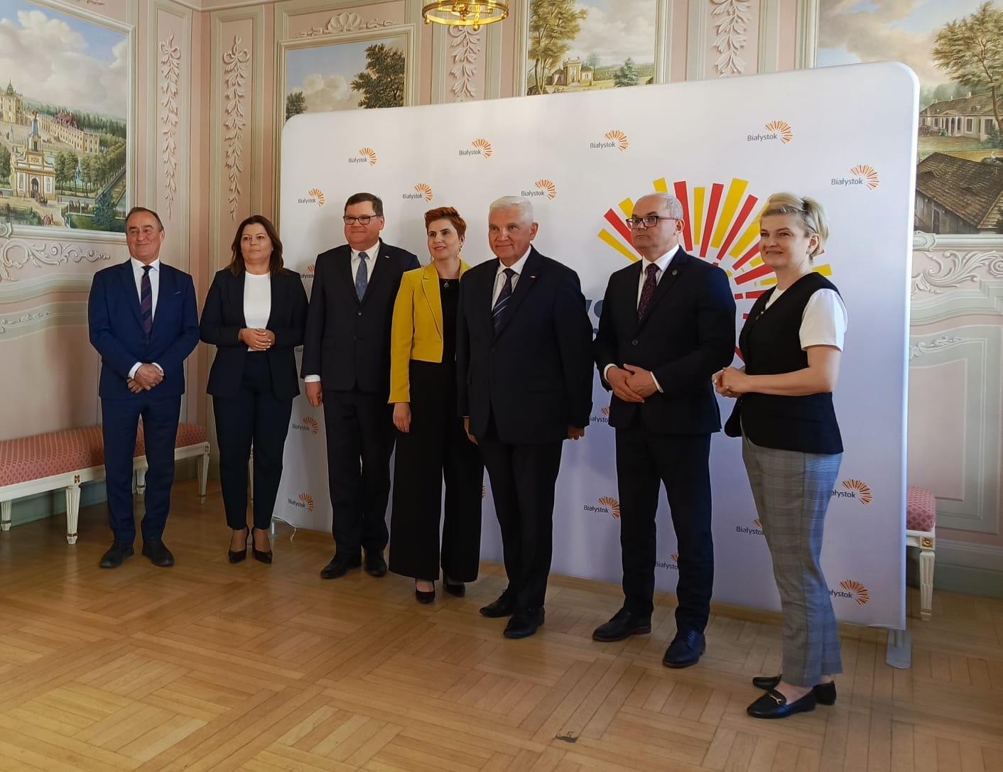 Grupa siedmiorga osób w eleganckich strojach w tym Prezydent Białegstoku na tle ścianki promującej miasto Białystok z kolorowym logo. Zdjęcie wykonane we wnętrzu ozdobnej sali Pałacyku Gościnnego w Białymstoku z malowidłami ściennymi i parkietem.