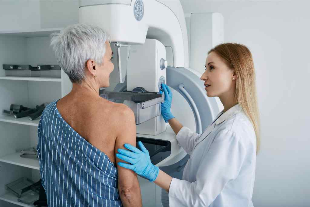 kobieta z siwymi wlosami poddaje sie mammografii obok lekarz mloda kobieta blond dlugie wlosy w rekawiczkach robi badanie w mammofrafie
