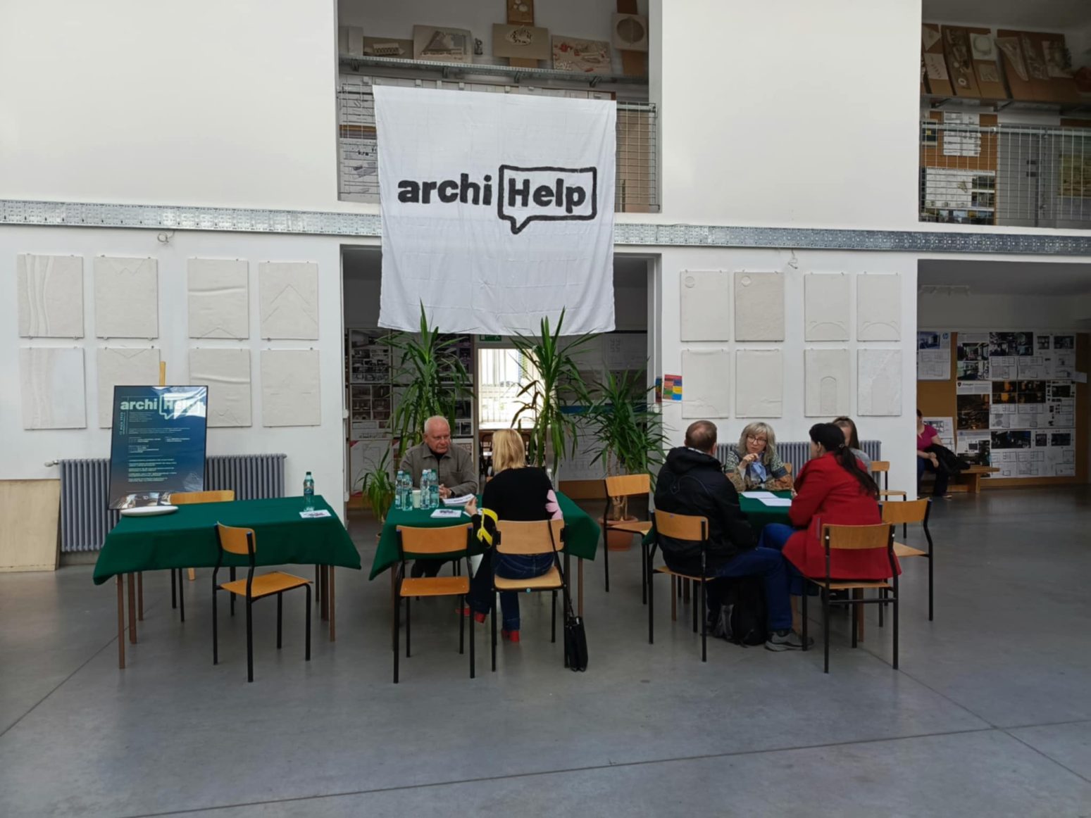 Na zdjęciu widać wydarzenie odbywające się w pomieszczeniu wewnętrznym, pod dużym banerem z napisem „archiHelp” w stylizowanej chmurce dialogowej, co sugeruje, że jest to inicjatywa związana z pomocą architektoniczną lub konsultacjami. Ludzie siedzą przy stołach, prawdopodobnie uczestnicząc w indywidualnych lub grupowych rozmowach bądź konsultacjach. Miejsce to wydział architektury PB — w tle widać modele architektoniczne i plakaty projektowe, co dodatkowo potwierdza architektoniczny charakter wydarzenia. Po lewej stronie znajduje się widoczna tablica z informacjami lub harmonogramem, również z napisem „archi”, prawdopodobnie prezentująca plan dnia lub oferowane usługi.