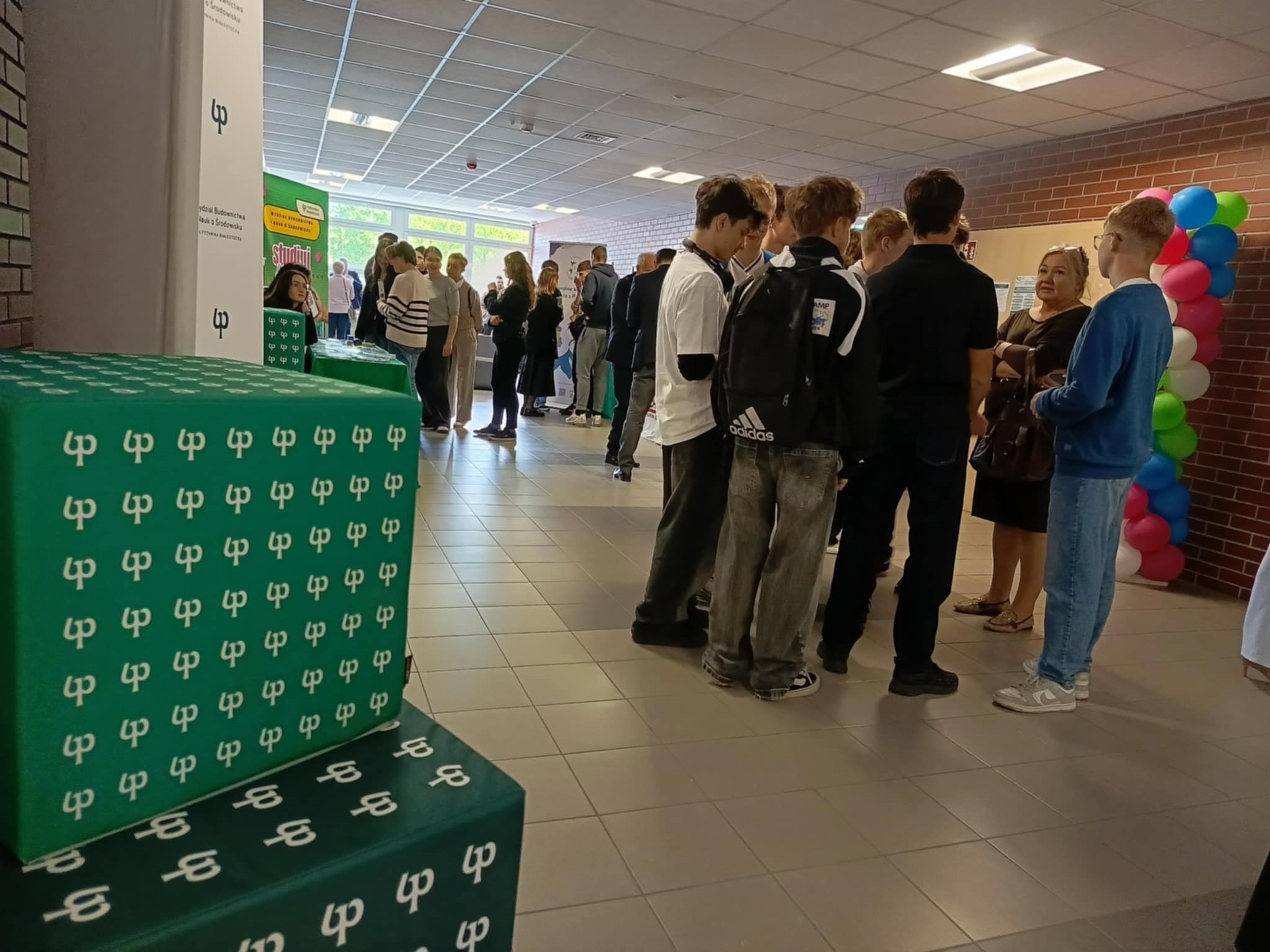 Korytarz uczelni stoja mlodzi ludzi, uczniowie, studenci, nauczyciele wykladowcy, na pierwszym planie widac logotypy Politechniki Bialostockiej, trwa przerwa przed wykladem