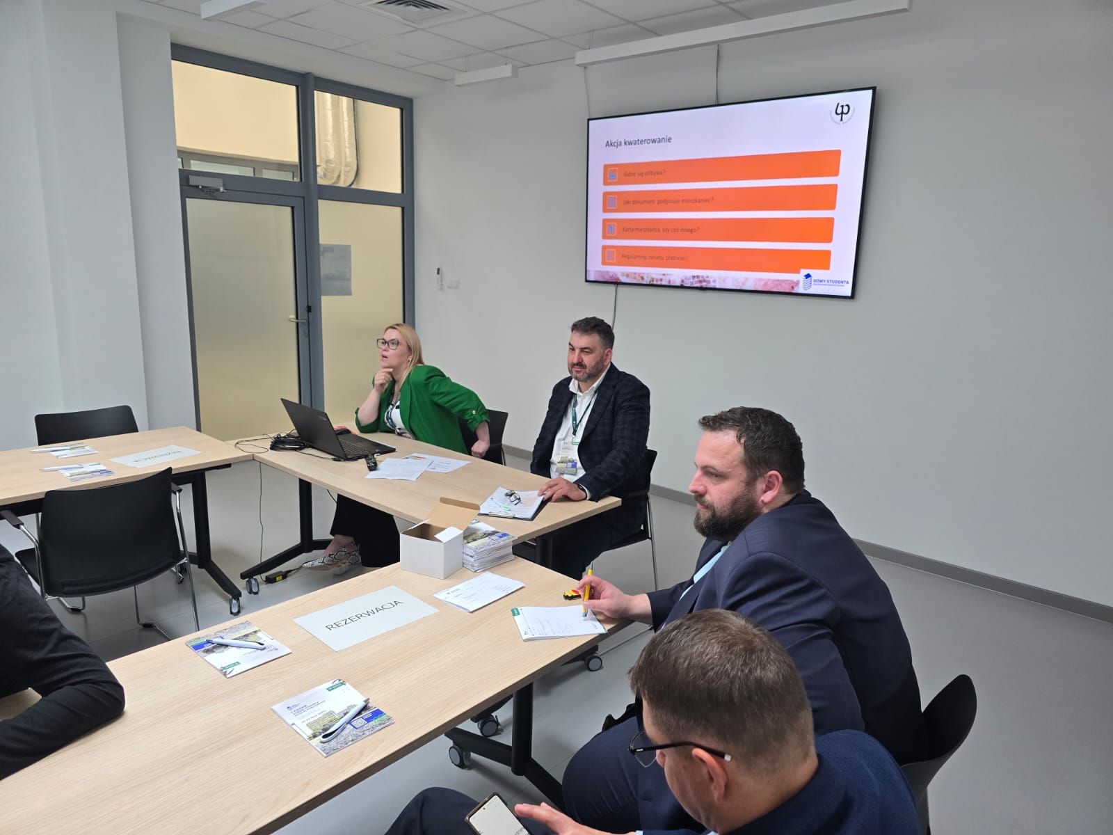 Prowadzący panel siedzą przy laptopie, w tle ekran z planszą „Akcja kwaterowanie” i punktami dotyczącymi zakwaterowania studentów