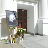 Zdjęcie miejsca pamięci przed Uniwersytetem Warszawskim: portret kobiety z czarną wstęgą, kwiaty i znicze ustawione przy wejściu do budynku z białymi kolumnami.