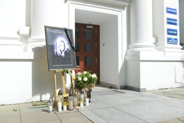 Zdjęcie miejsca pamięci przed Uniwersytetem Warszawskim: portret kobiety z czarną wstęgą, kwiaty i znicze ustawione przy wejściu do budynku z białymi kolumnami.