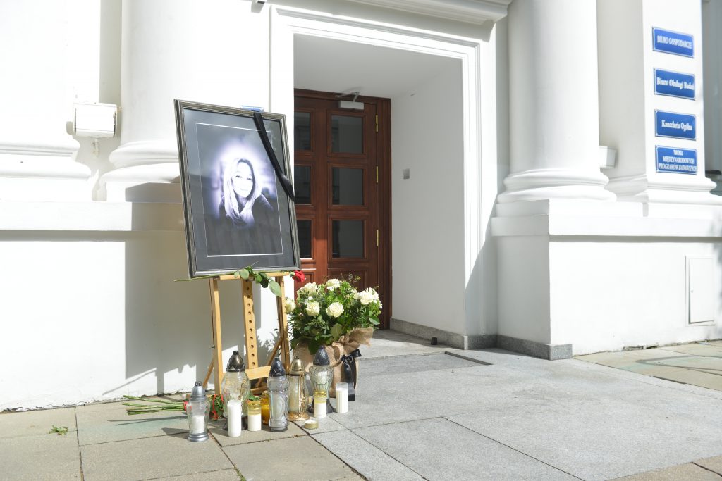 Zdjęcie miejsca pamięci przed Uniwersytetem Warszawskim: portret kobiety z czarną wstęgą, kwiaty i znicze ustawione przy wejściu do budynku z białymi kolumnami.