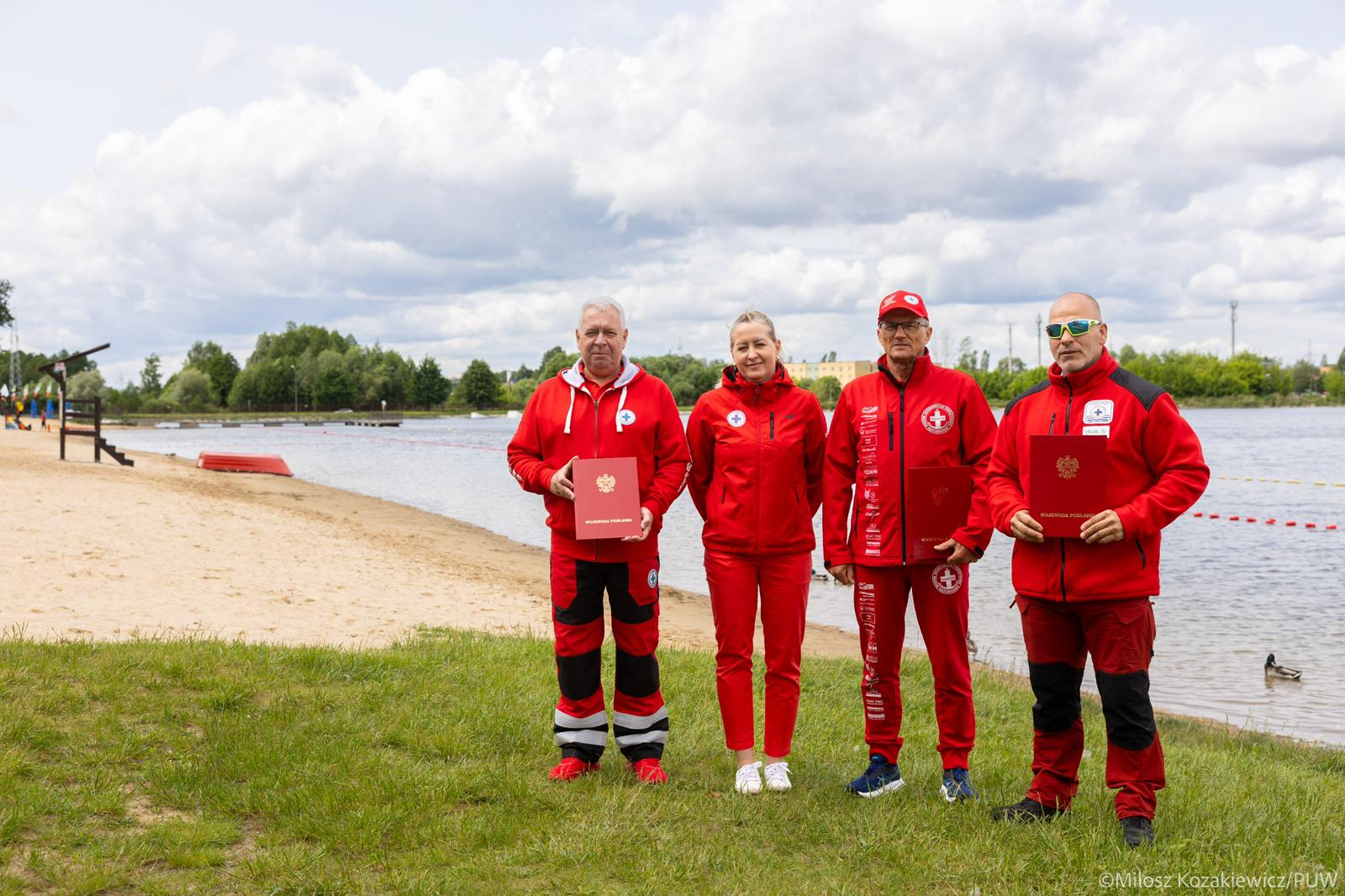 Na zdjęciu widzimy czworo ratowników wodnych ubranych w czerwone stroje, stojących na tle plaży i zbiornika wodnego. Troje z nich trzyma w rękach czerwone teczki z godłem Rzeczypospolitej Polskiej – najprawdopodobniej są to wyróżnienia lub podziękowania za służbę. Ich profesjonalna postawa i sceneria wskazują na oficjalne wydarzenie związane z bezpieczeństwem nad wodą lub początkiem sezonu kąpielowego.