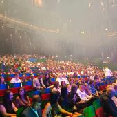 Zdjęcie przedstawia salę wypełnioną publicznością, najprawdopodobniej podczas gali lub dużego wydarzenia artystycznego. Widać kolorowe, numerowane fotele, nowoczesną salę widowiskową z dwoma poziomami oraz sceniczne oświetlenie i lekki dym sceniczny – co sugeruje, że wydarzenie już trwa lub zaraz się rozpocznie. Publiczność jest różnorodna – młodzież, dorośli, osoby starsze – co świadczy o szerokim zainteresowaniu wydarzeniem. Atmosfera jest uroczysta i pełna oczekiwania. Jeśli chcesz, mogę napisać relację lub opis wydarzenia do publikacji.