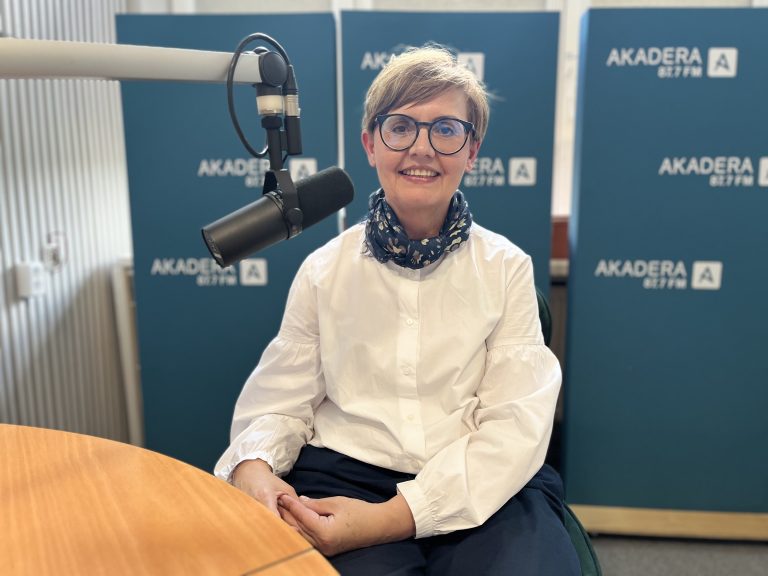 Monika Kordiukiewicz siedzi w studiu Radia Akadera przy mikrofonie, na tle niebieskich plansz z logo stacji. Ma krótkie jasne włosy, okulary i biały koszulowy top.