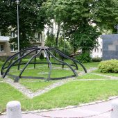 Pomnik upamiętniający Wielką Synagogę w Białymstoku – metalowa konstrukcja w kształcie kopuły oraz czarna płyta z menorą, ustawione na trawiastym skwerze wśród drzew i otaczających bloków.